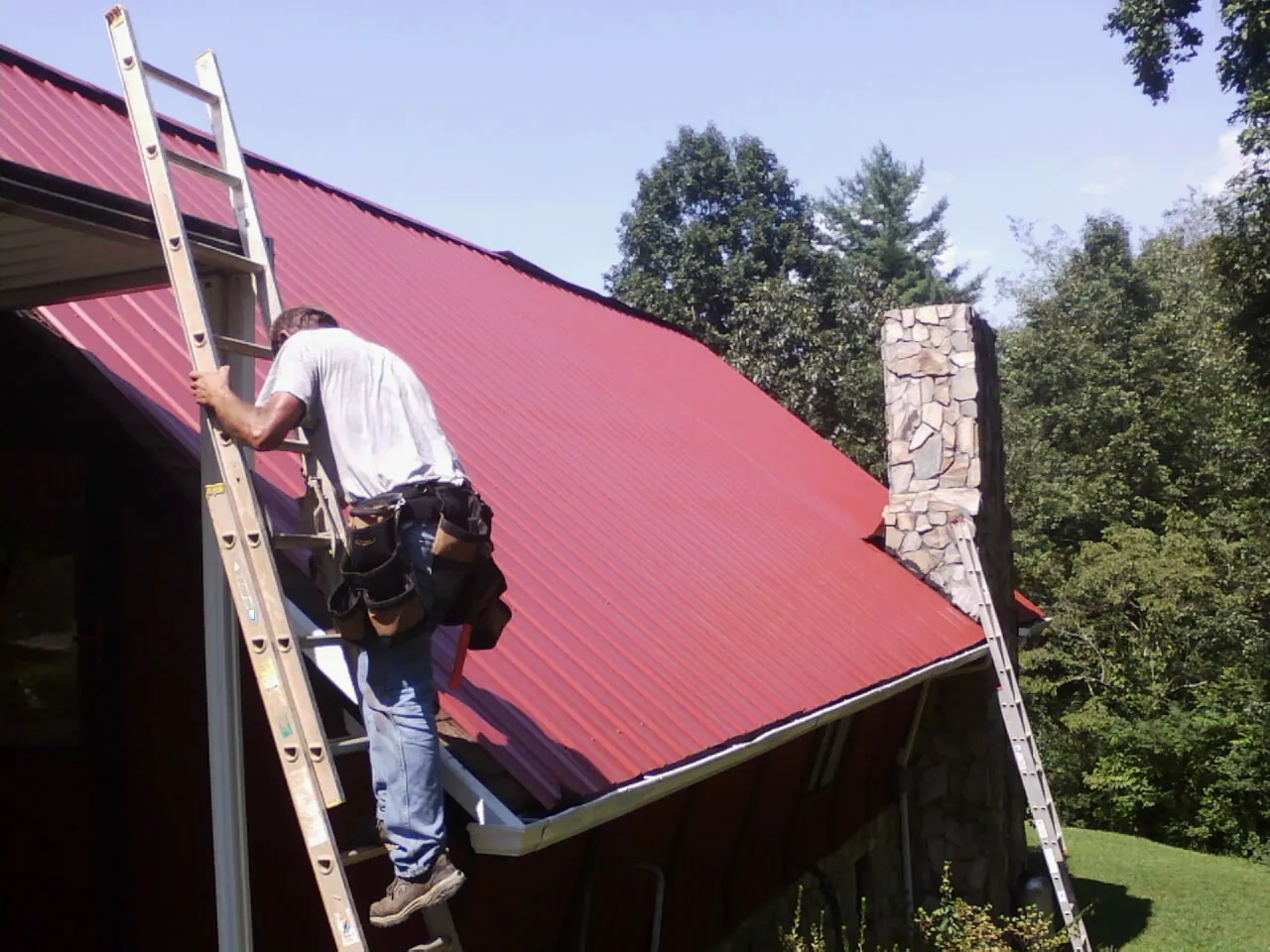Roofer on a ladder beside a red metal roof and stone chimney — Soffit & Fascia Repair by Roofing Brothers in Josephine